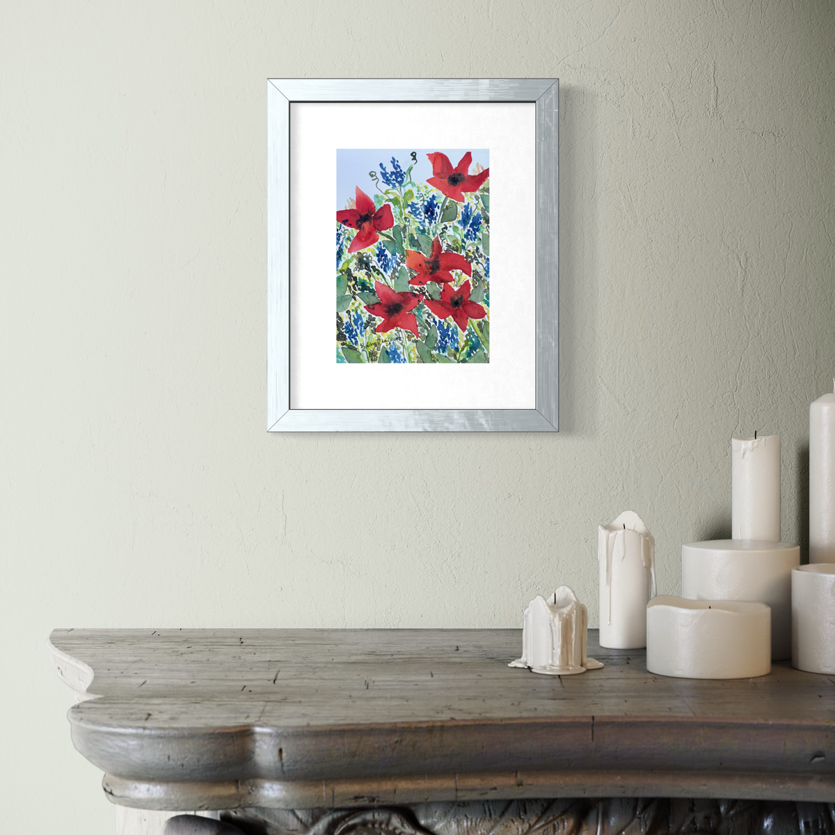 A framed floral art painting featuring red poppies and blue flowers on a light blue background, displayed on a textured beige wall above a rustic wooden mantel with white candles.