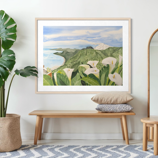 Framed artwork of calla lilies and mountains in a room with a bench and plant.