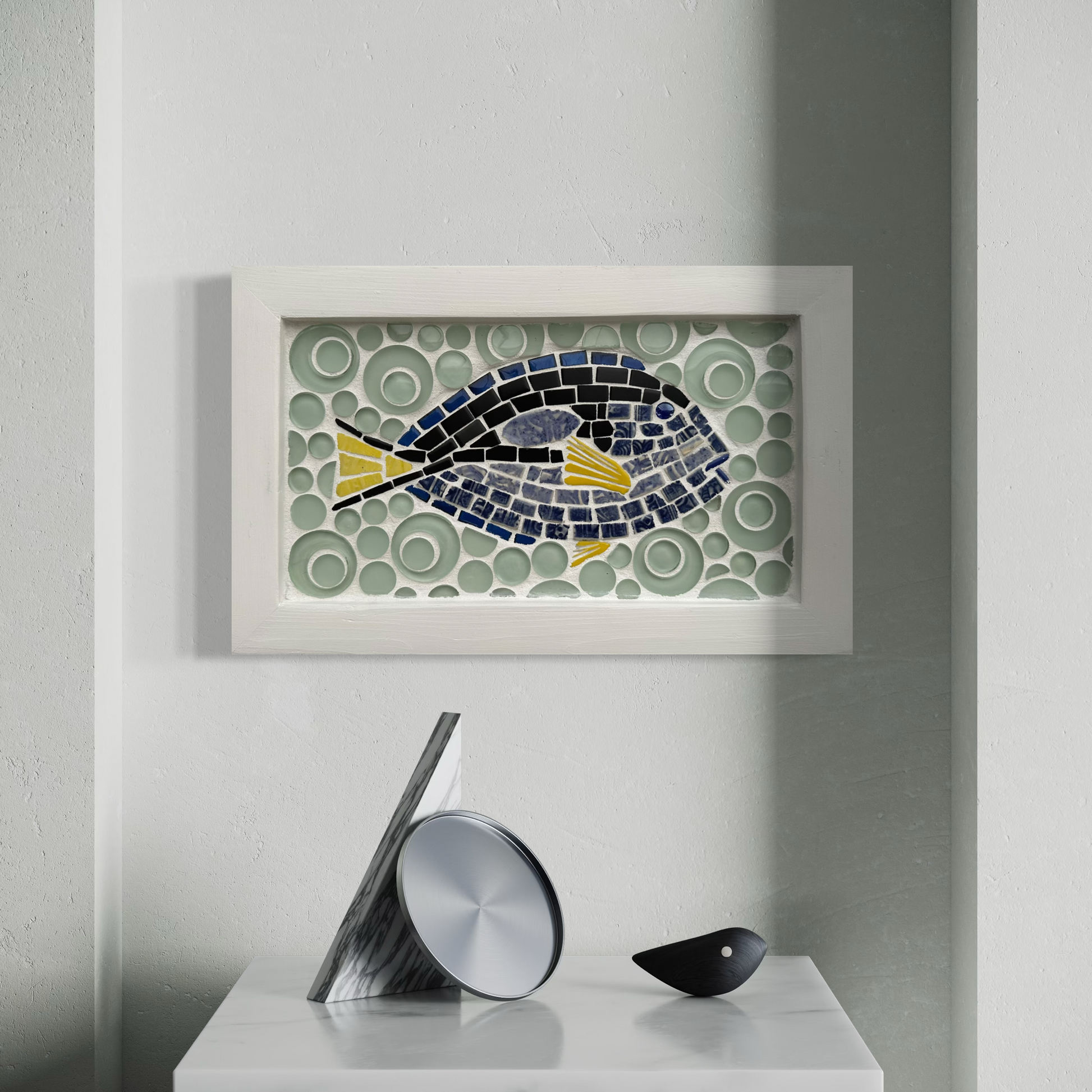Framed mosaic artwork of a fish on a wall with decorative objects below.