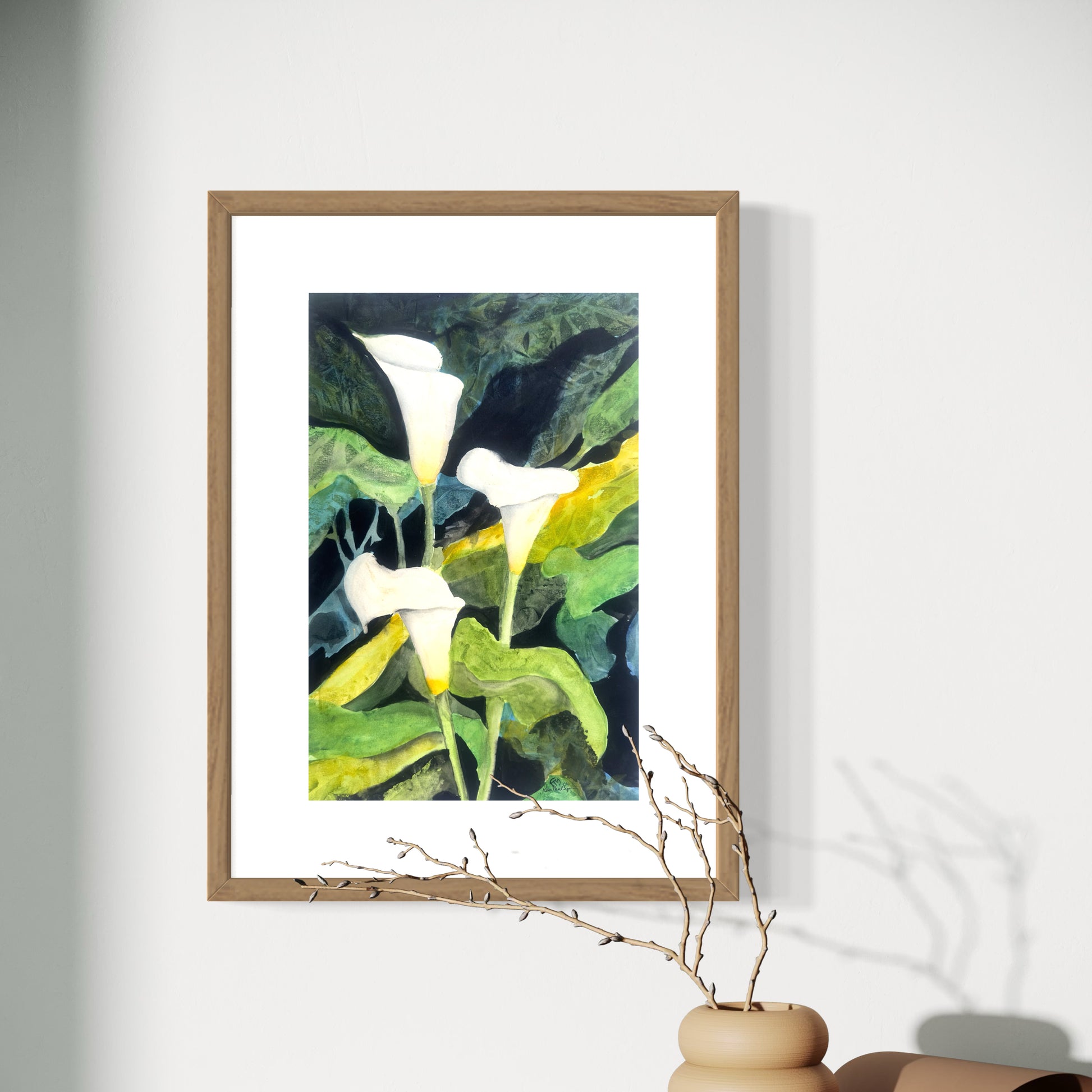 Framed watercolor artwork of 3 calla lilies on a white wall with a vase and branches in the foreground.