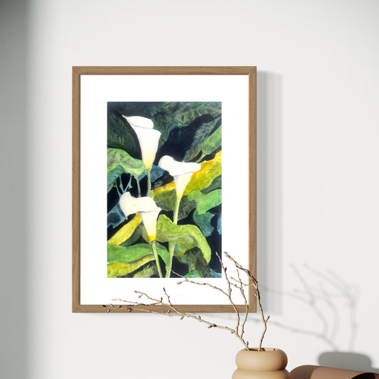 Framed watercolor artwork of 3 calla lilies on a white wall with a vase and branches in the foreground.