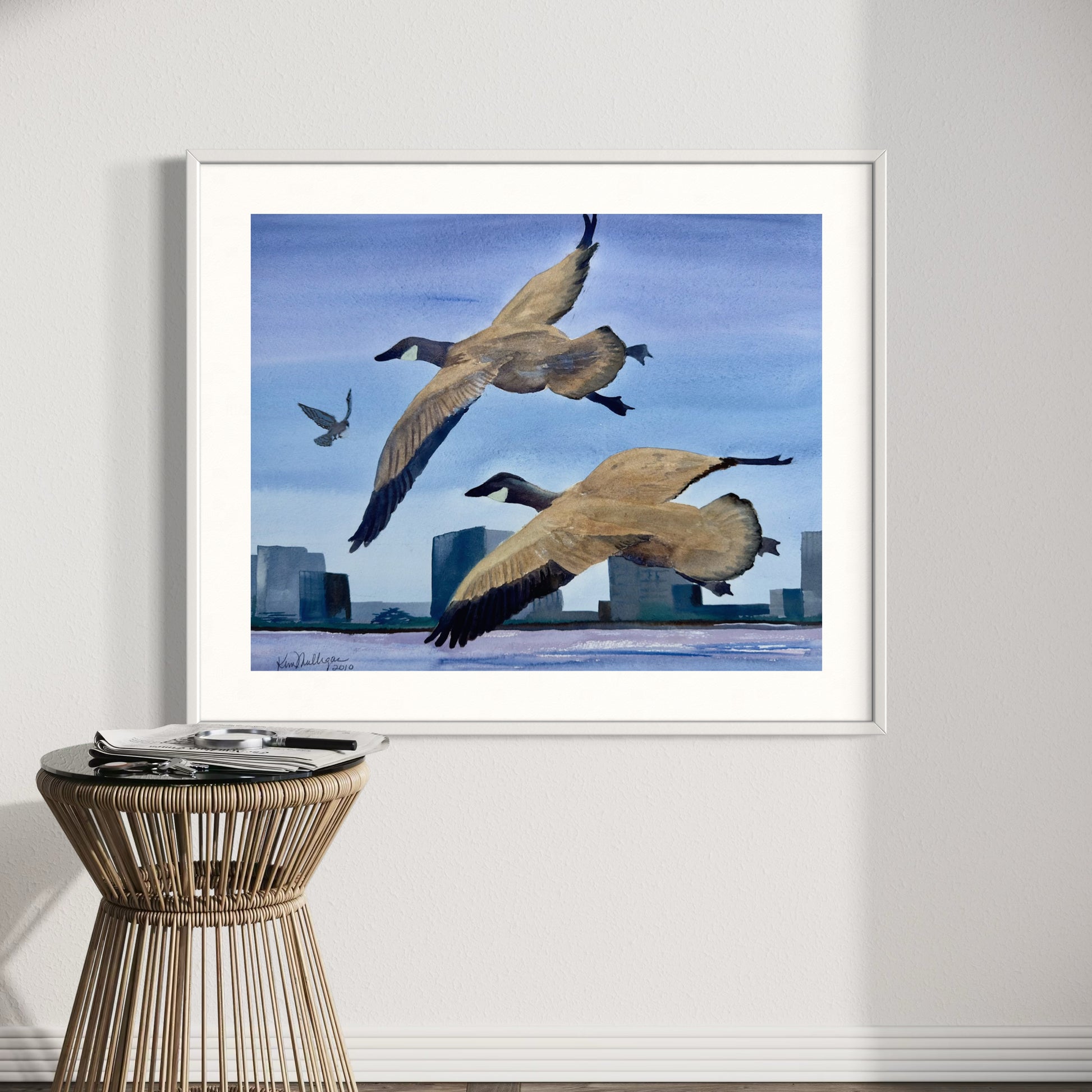 Framed artwork of geese flying over a cityscape on a wall above a small table.