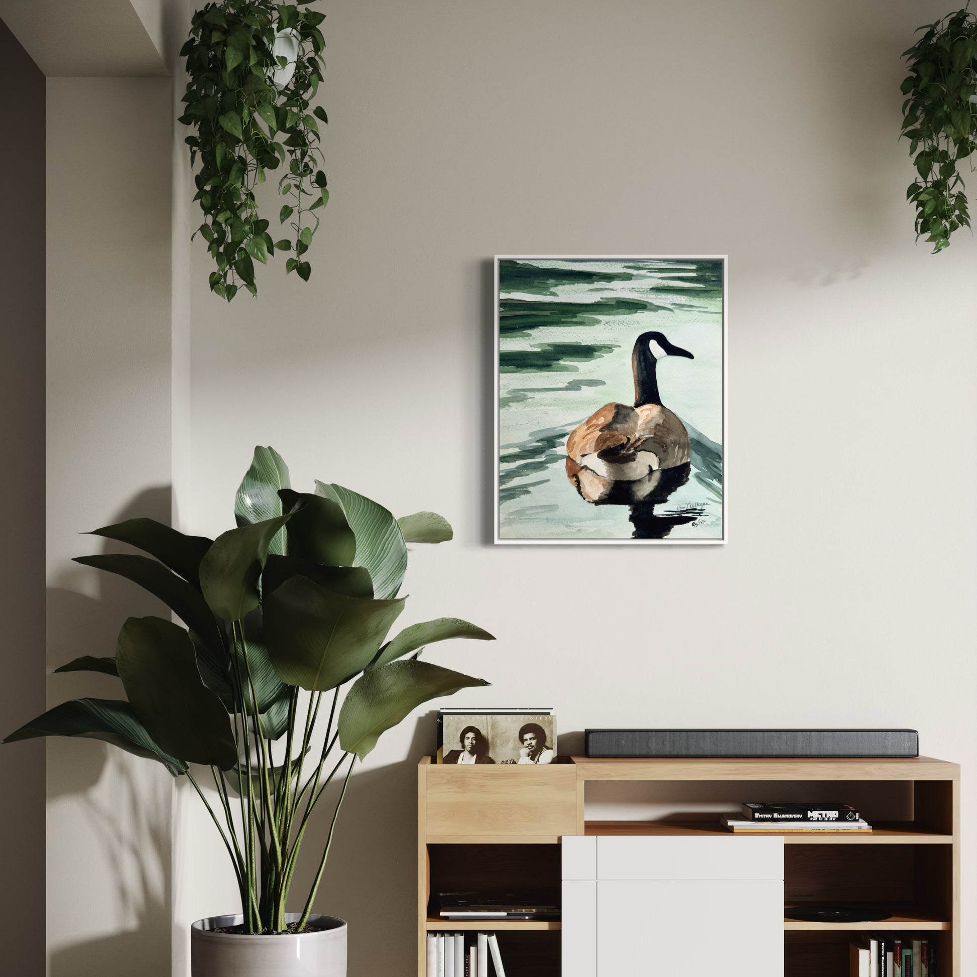 Framed watercolor artwork of a Canada goose on a wall above a wooden shelf with plants and decor.