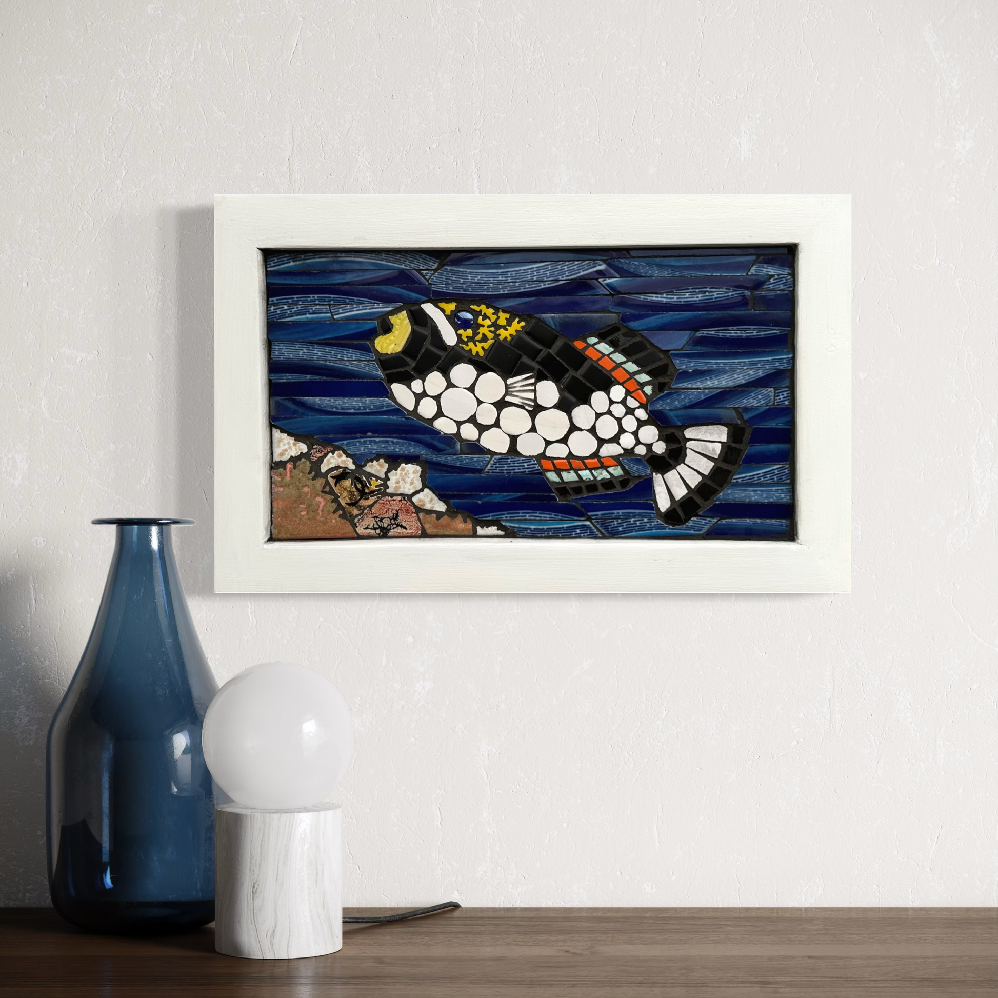 Framed mosaic artwork of a fish on a wall with a blue vase and white lamp below.