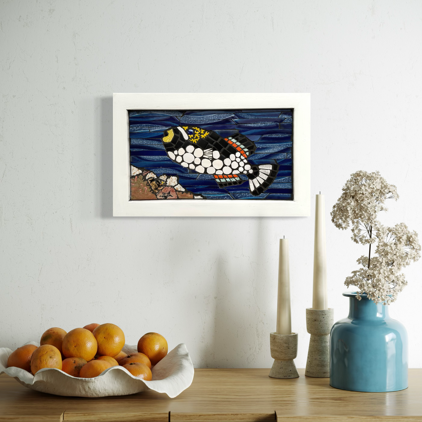 Framed artwork of a mosaic fish on a wall above a table with fruit, candles, and a vase.