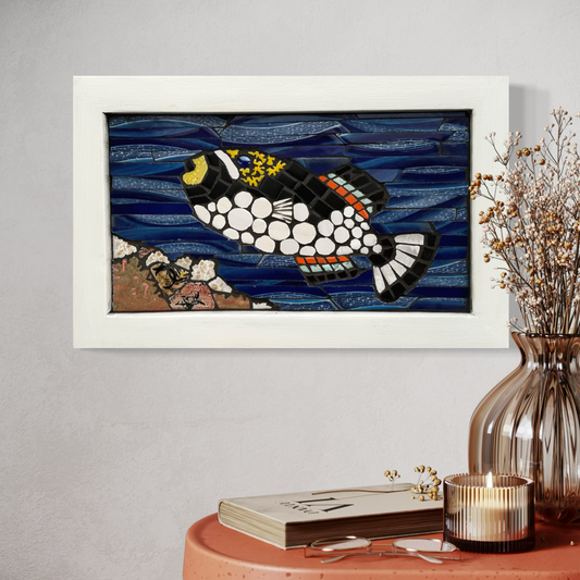 Framed mosaic artwork of a clown triggerfish on a wall with decorative items on a table below.