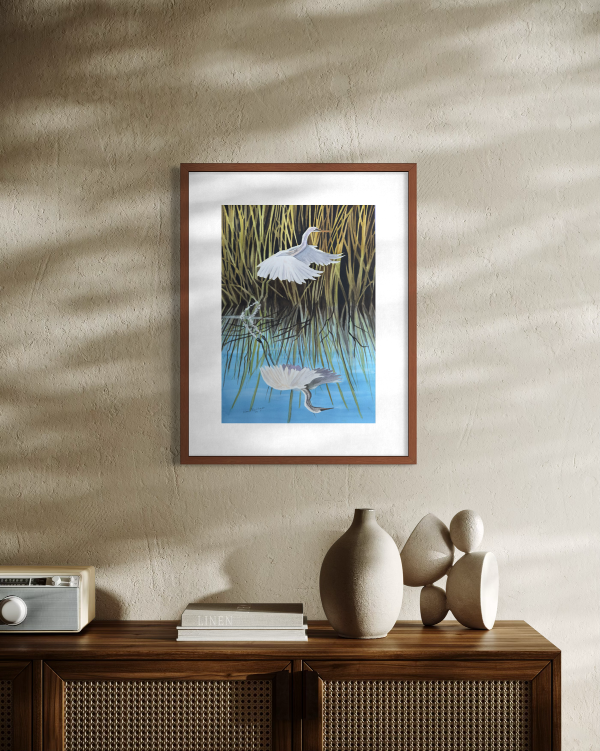 A framed art depicting a white bird in a marsh under blue water, styled on a textured beige wall above a wooden cabinet with decorative ceramic vases.
