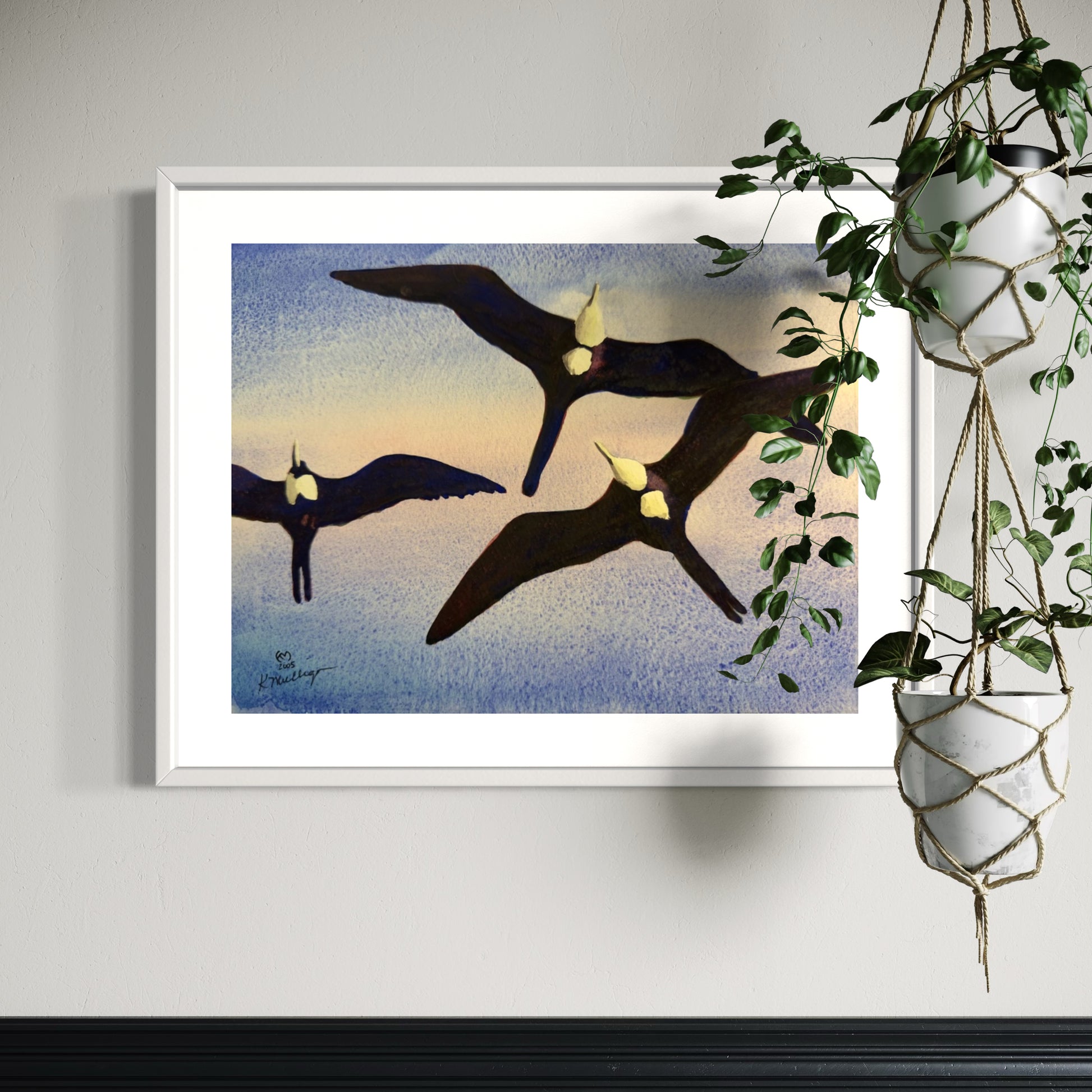 Framed artwork of birds flying against a sunset sky, hanging on a wall with a plant to the right.