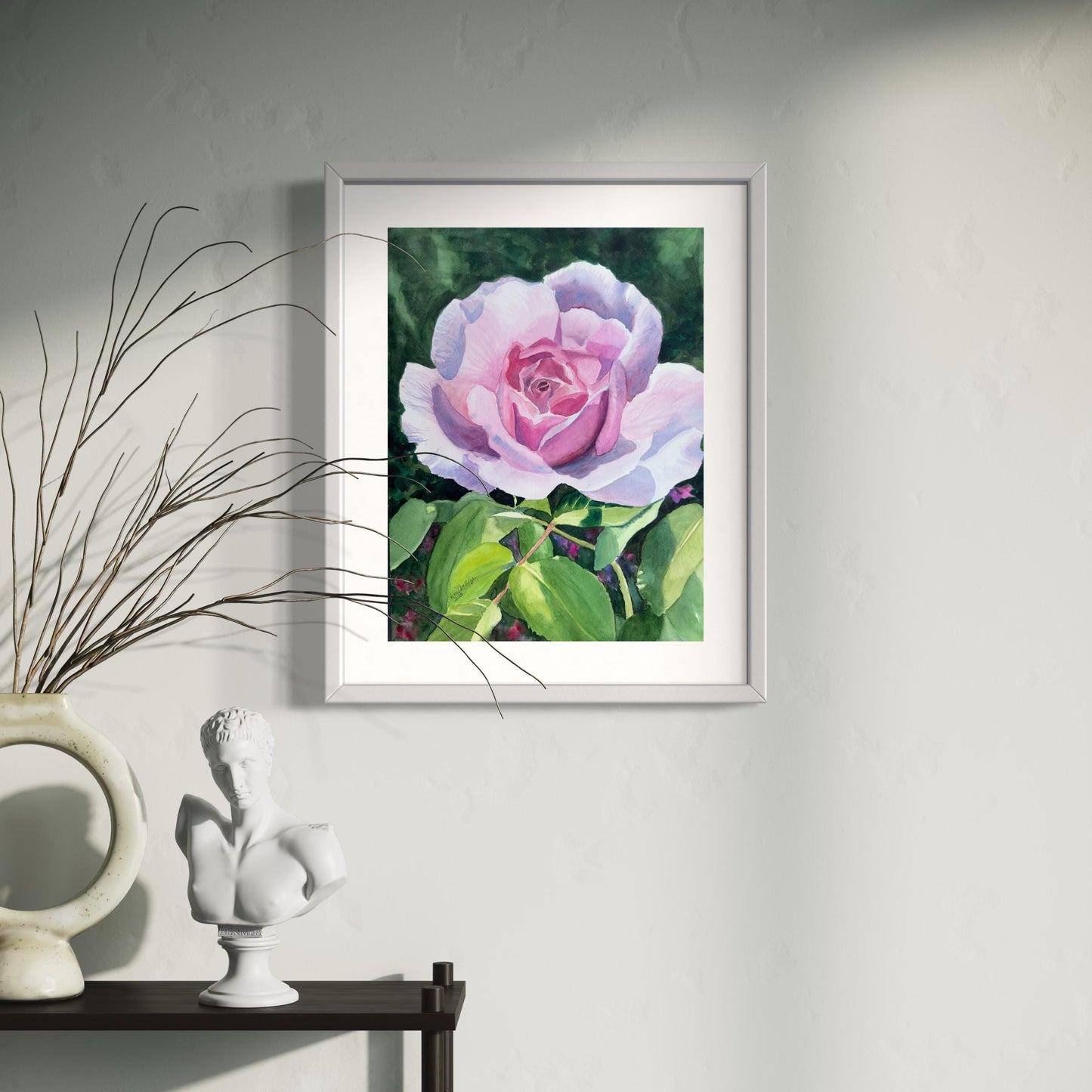 Framed watercolor artwork of a pink rose on a wall with decorative elements below.