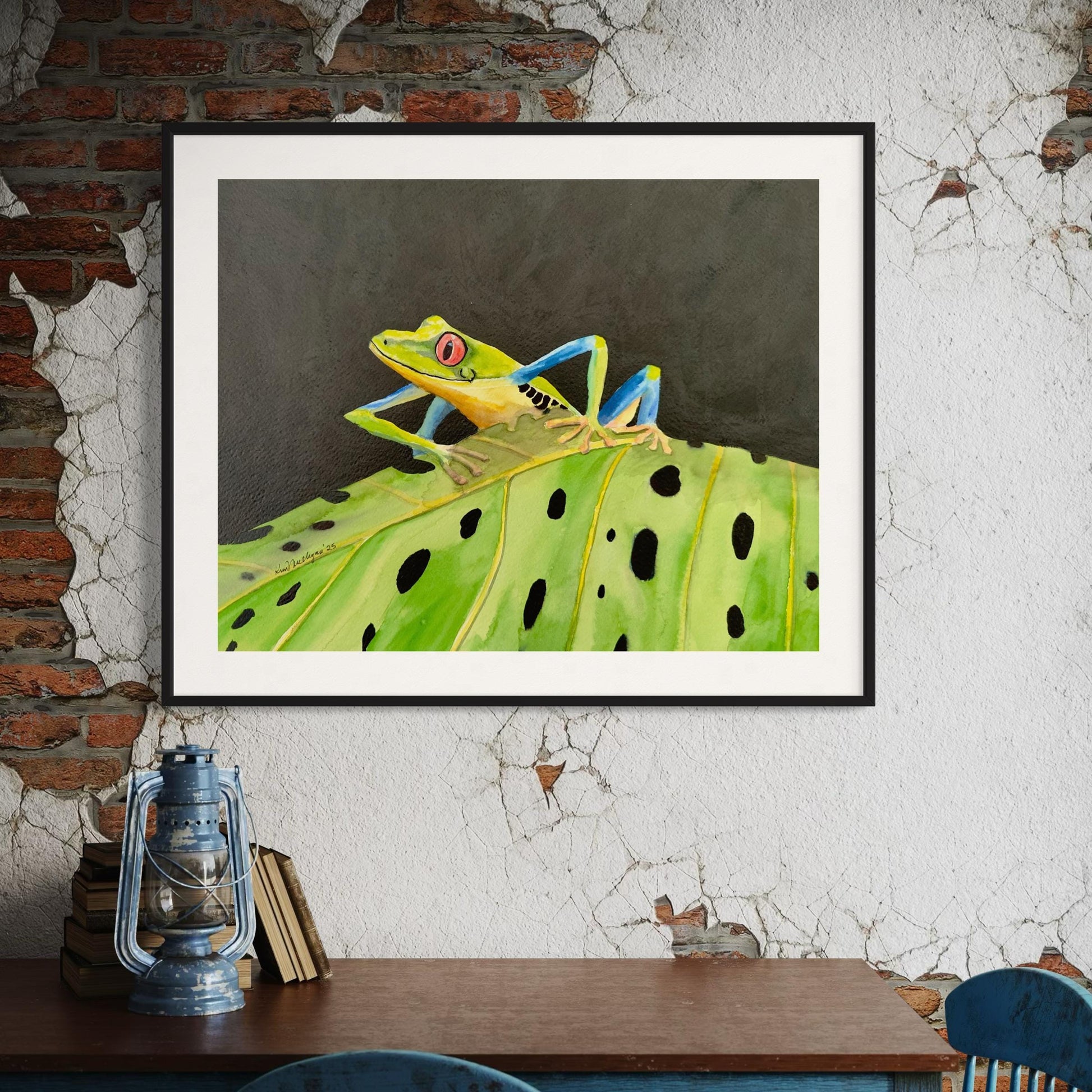 Framed watercolor artwork of a red eyed tree frog on a leaf against a textured wall.