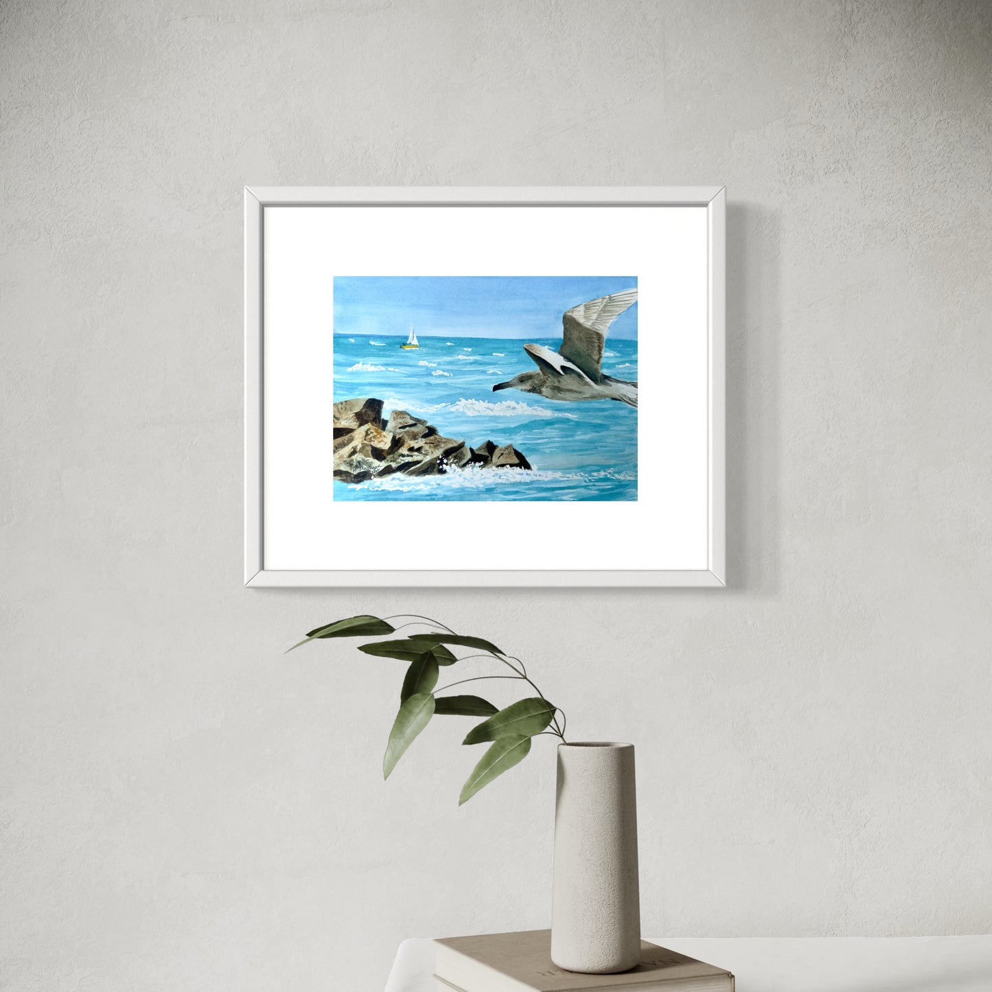 Framed watercolor artwork of a seagull over water with rocks, displayed on a wall above a white vase with green leaves.