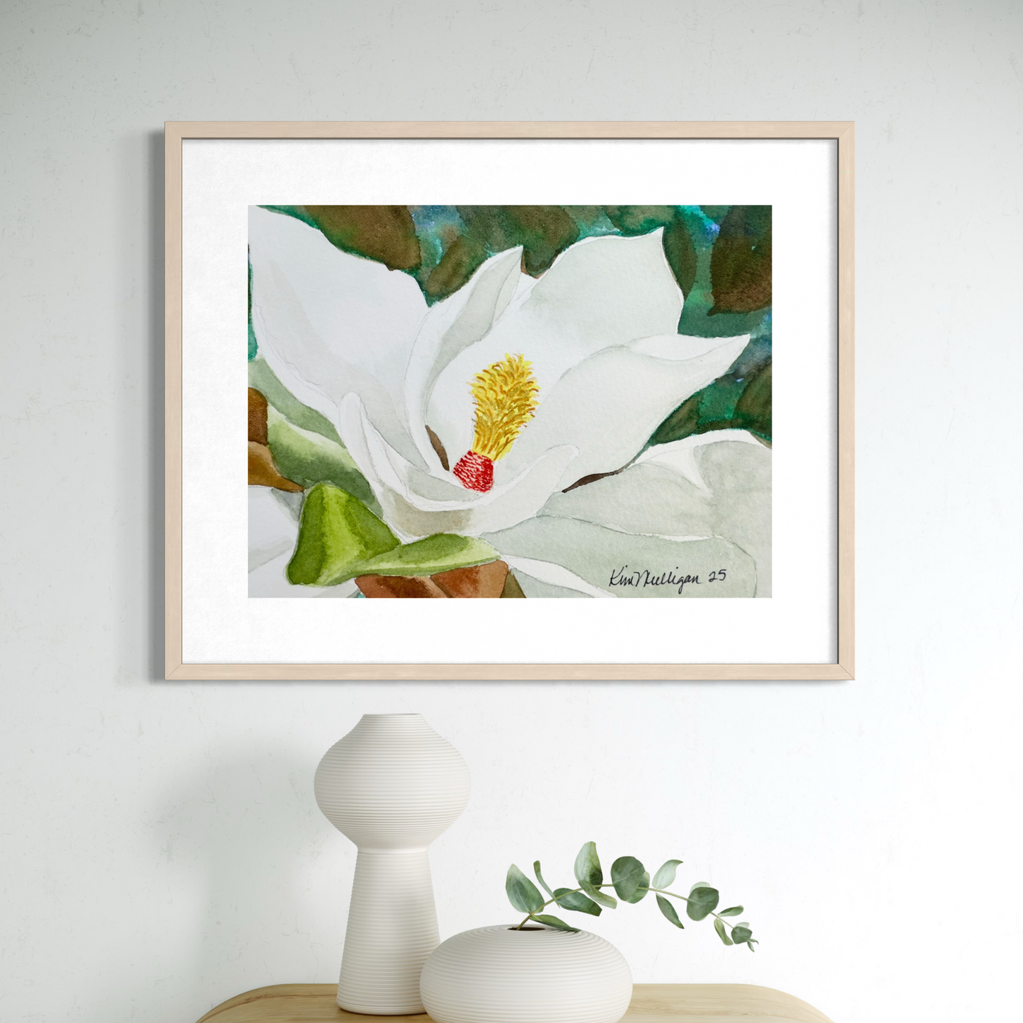 Framed watercolor artwork of a white magnolia flower with green leaves on a light gray wall.