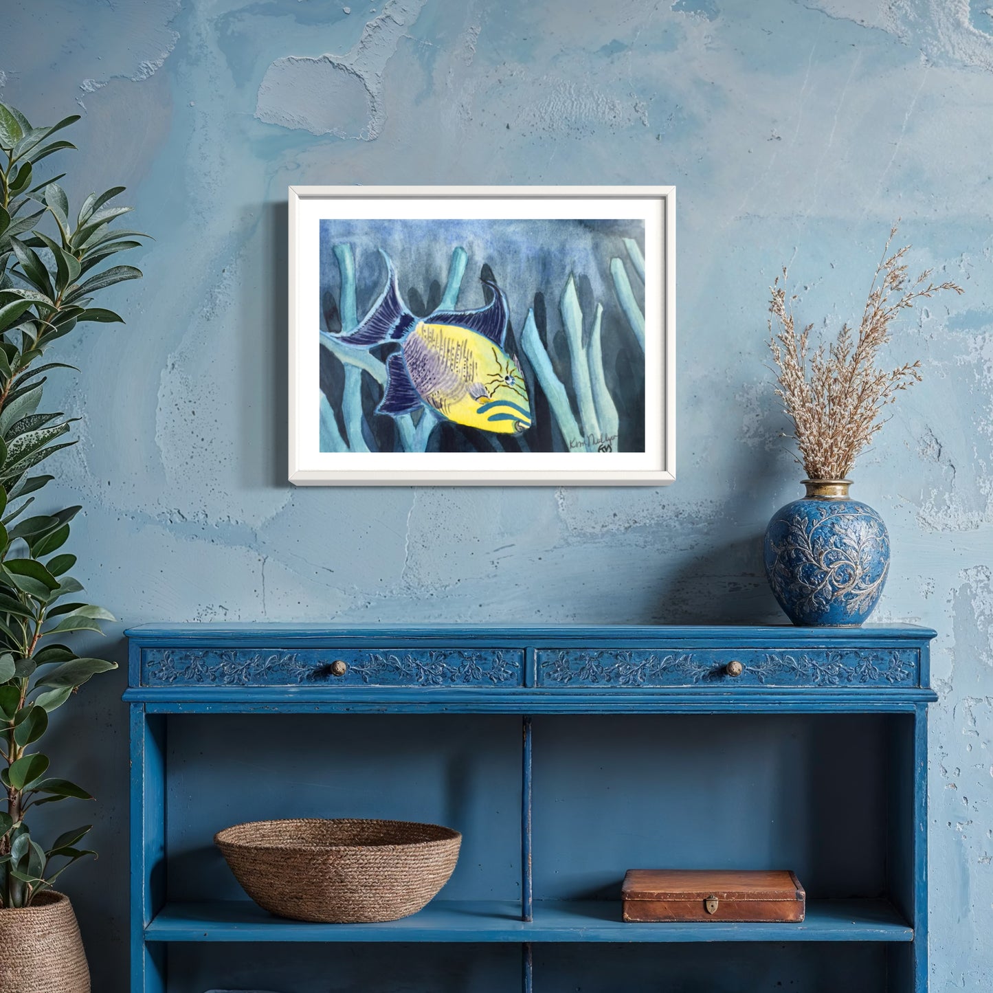 Framed watercolor artwork of queen trigger fish on a blue wall above a blue shelf with decorative items.