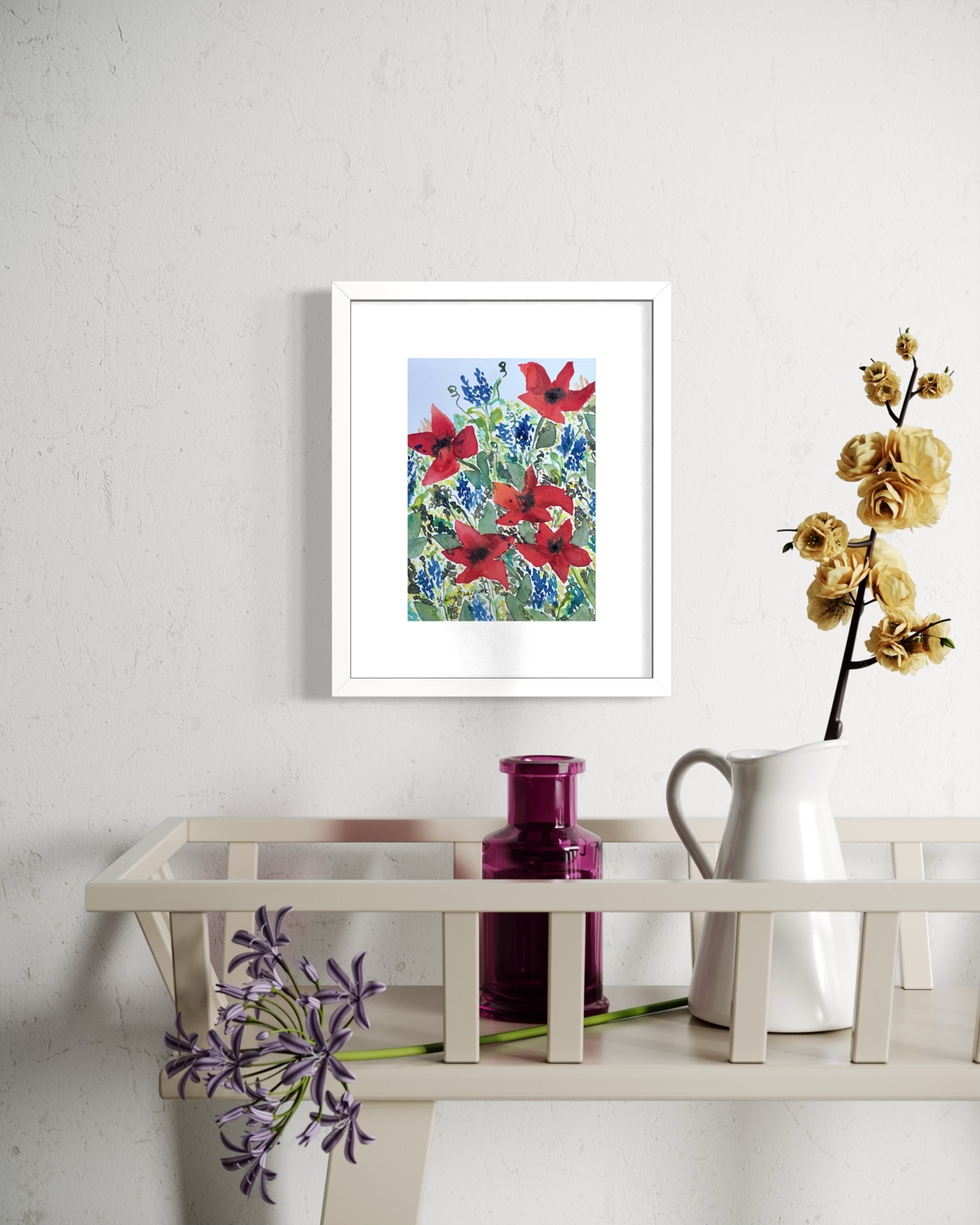 A framed floral art print featuring red poppies and blue flowers hangs on a white textured wall above a light wood console table styled with a white pitcher, a purple bottle, and purple flowers.