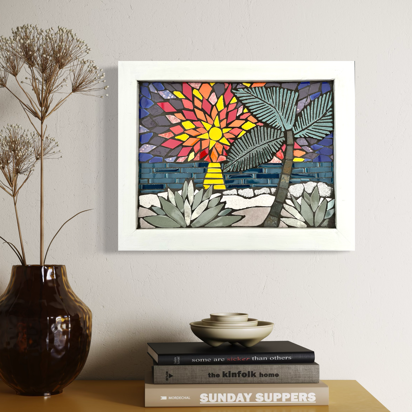 Framed abstract mosaic artwork on a wall above a yellow table with books and a vase.