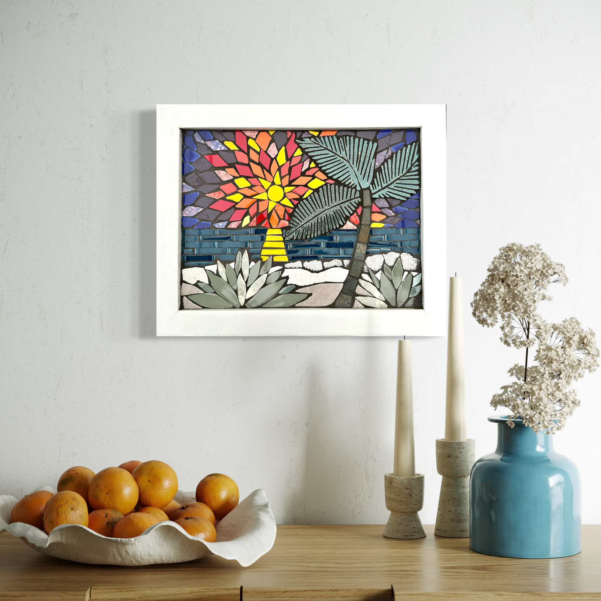 Framed colorful mosaic seascape artwork on a wall above a table with oranges, candles, and a vase.