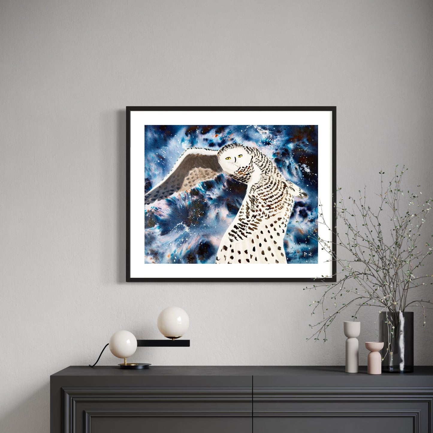 Framed watercolor artwork of a snowy owl against a cosmic sky on a gray wall above a black cabinet.