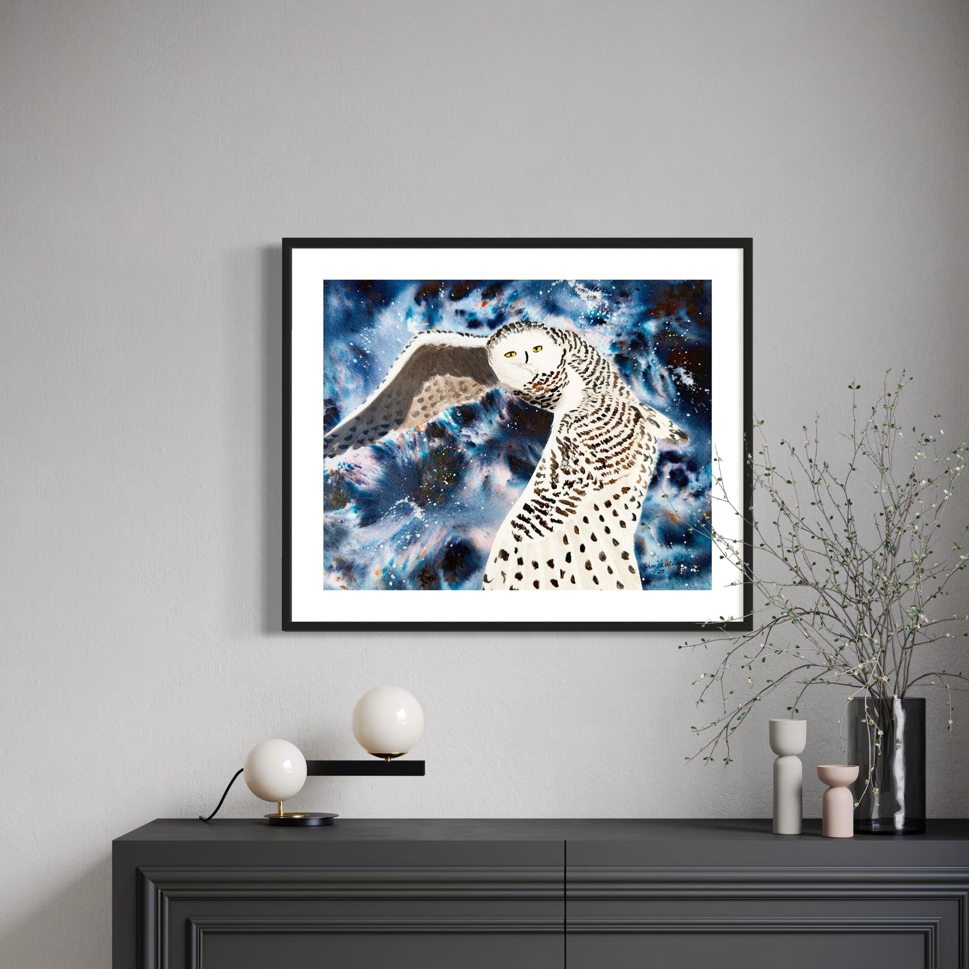 Framed watercolor artwork of a snowy owl against a cosmic sky on a gray wall above a black cabinet.