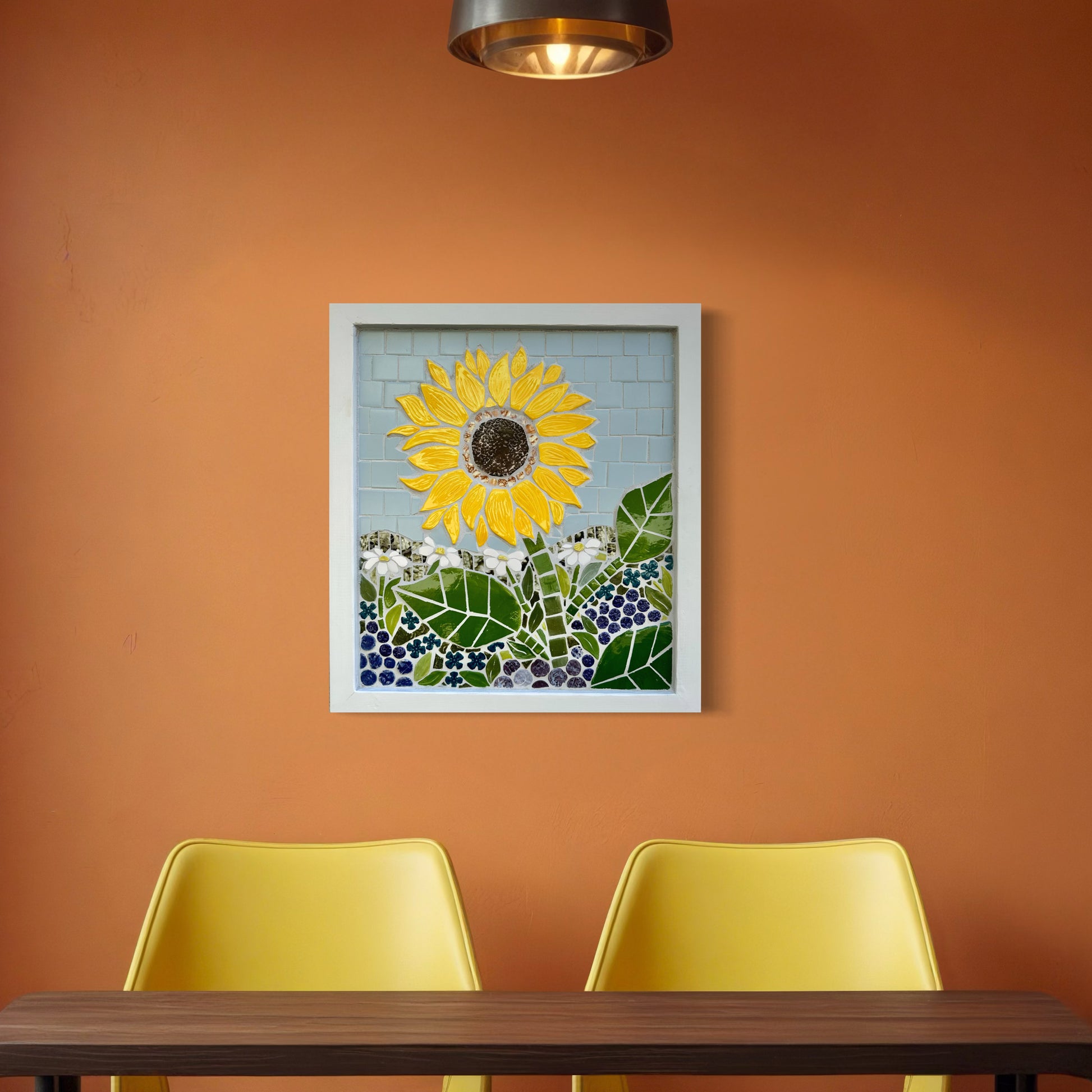 Framed mosaic artwork of a yellow sunflower on an orange wall with two yellow chairs and a wooden table.