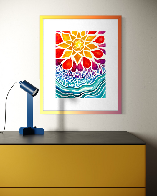 A framed art print featuring a colorful watercolor sunflower and wave design, displayed on a wall above a yellow cabinet with a blue lamp in the foreground.