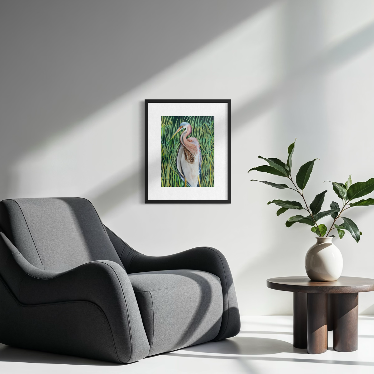A framed art print of a heron in a marsh is displayed on a light gray wall in a modern living room setting, next to a gray armchair and a small side table with a potted plant.