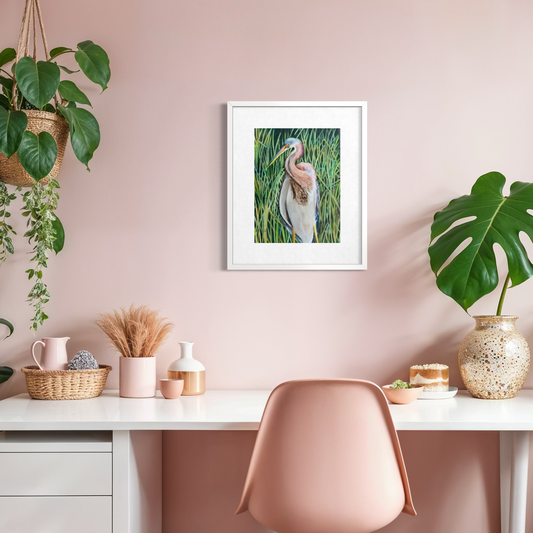 A framed watercolor painting of a tricolor heron in a marsh, displayed on a dusty pink wall above a white desk with a pink chair and decorative plants.