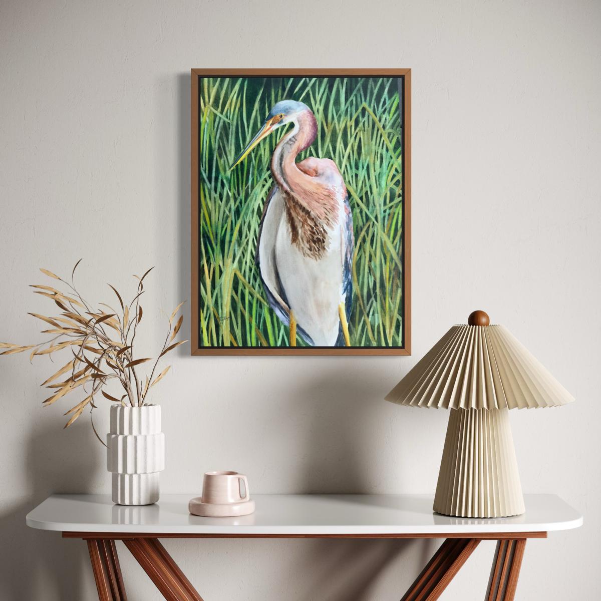 A framed painting of a heron in tall green reeds hangs on a light gray wall above a console table styled with a pleated beige table lamp, a white vase with dried branches, and a small pink cup.