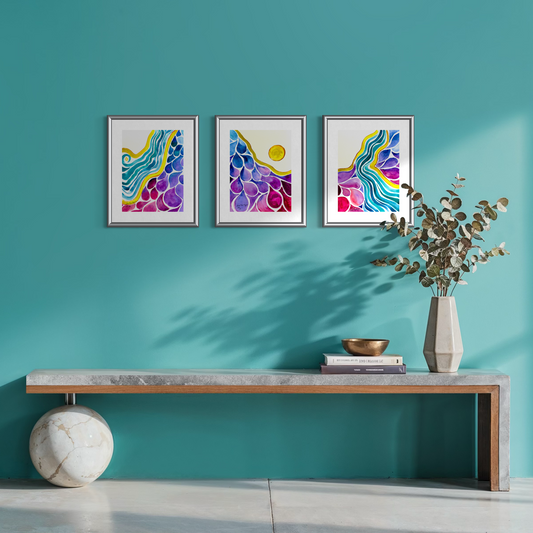 Three framed abstract paintings with colorful wave and bubble designs hang on a teal wall above a long console table with a vase of eucalyptus and decorative objects.